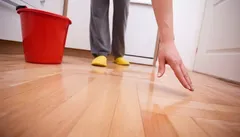 Persoon maakt houten vloer schoon met handdoek, rode emmer en gele pantoffels zichtbaar.