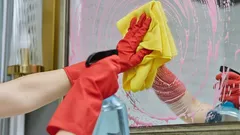 Persoon met rode handschoenen reinigt een spiegel met een gele doek en schoonmaakmiddel.