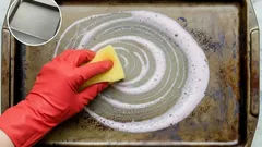 Vuile bakplaat schoonmaken met spons en reinigingsmiddel, hand met rode handschoen.
