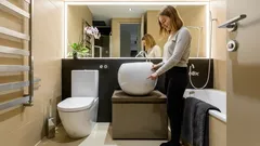 Vrouw in moderne badkamer bij waskom en spiegel, strakke inrichting en neutrale kleuren.